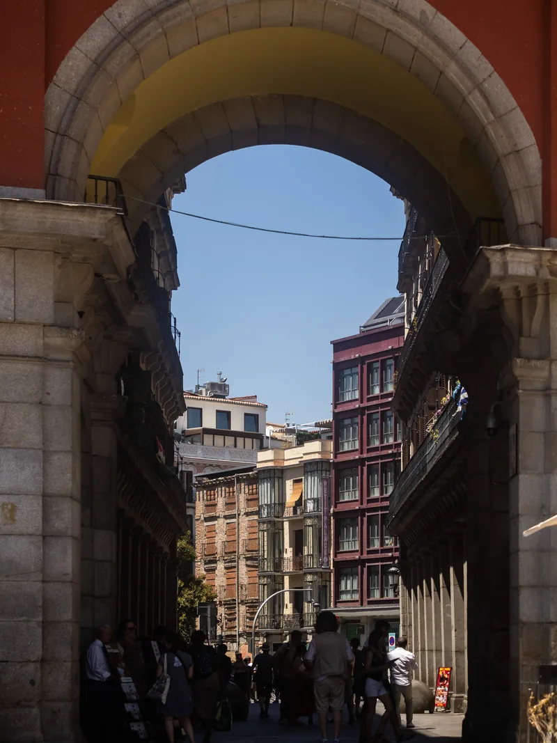 A street of tall buildings seen through a deep stone arch, with people walking in the shade.