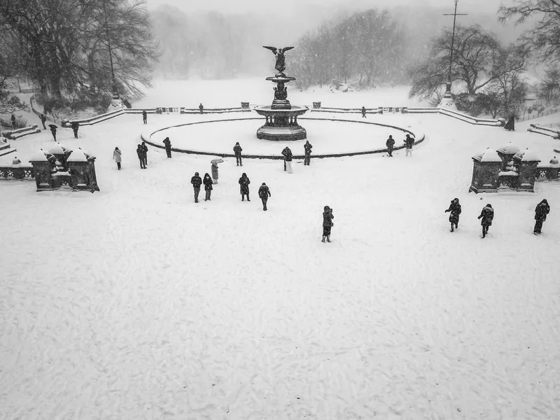 People walking across the snow-covered plaza around Bethesda Fountain.