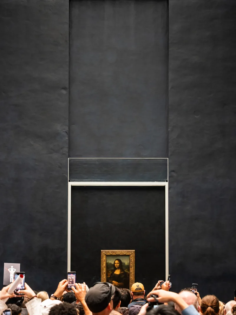 A crowd holding up phones toward the Mona Lisa, small against a tall dark wall.