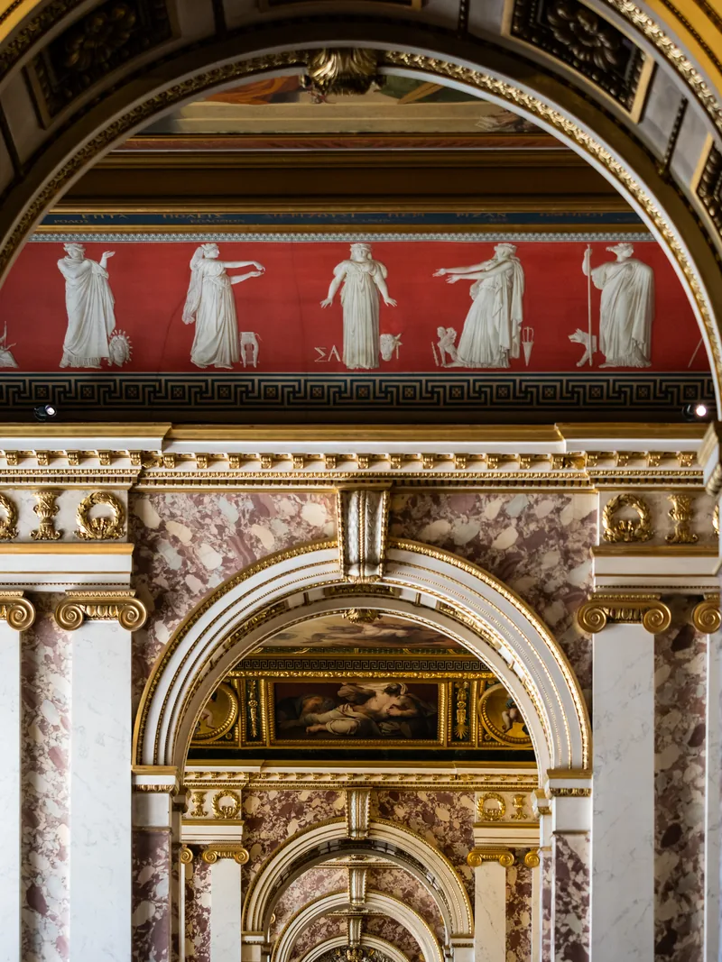 Gilded arches, marble columns and a red frieze of classical figures in a palace hallway at Versailles.