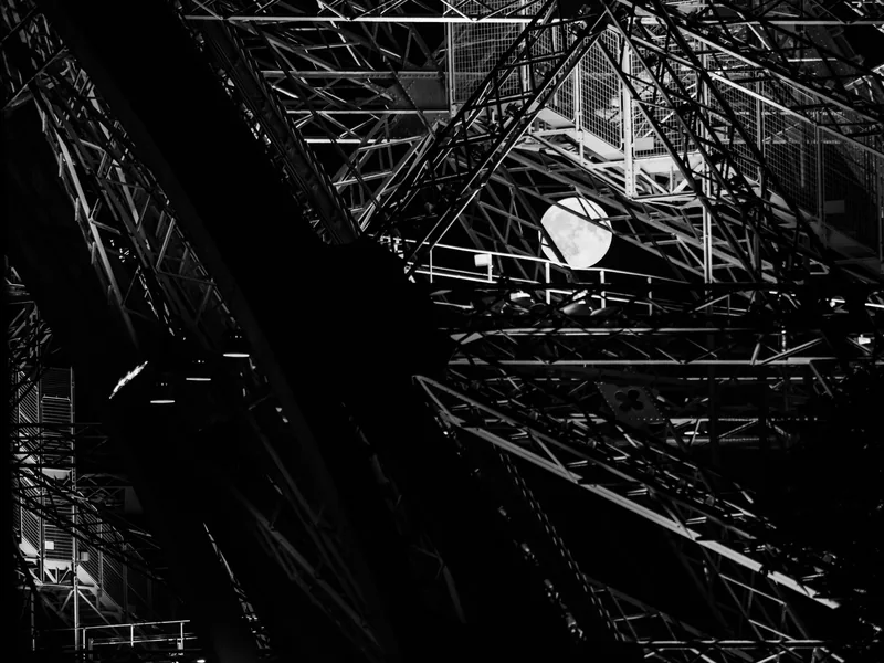 The Eiffel Tower's iron lattice seen from below in stark black and white, with the sun behind it.