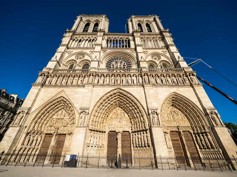 The west front of Notre Dame against a deep blue sky, with a crane arm at the edge of the frame.