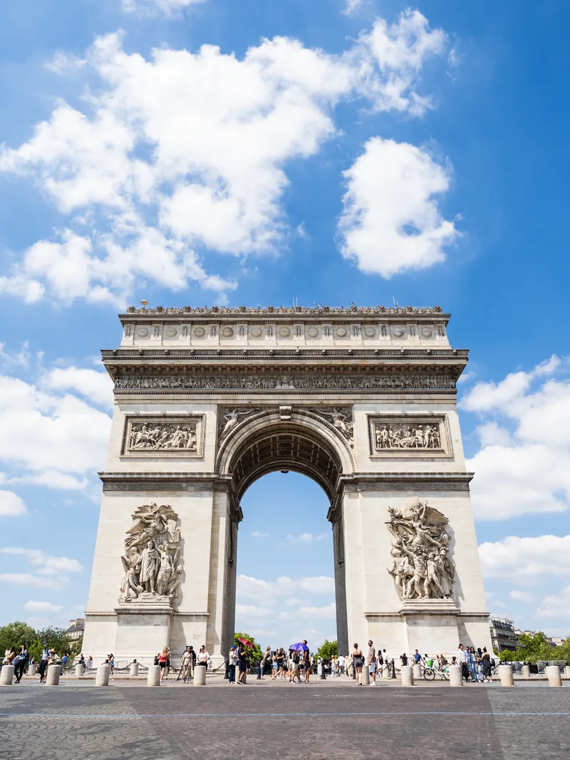The Arc de Triomphe seen from street level beneath puffy white clouds, with crowds at its base.