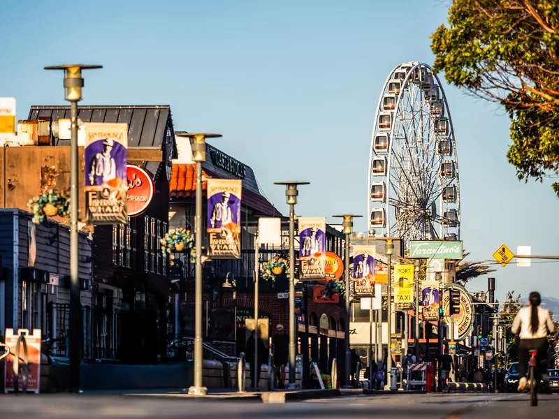 A waterfront street with banners and a Ferris wheel under a blue sky.