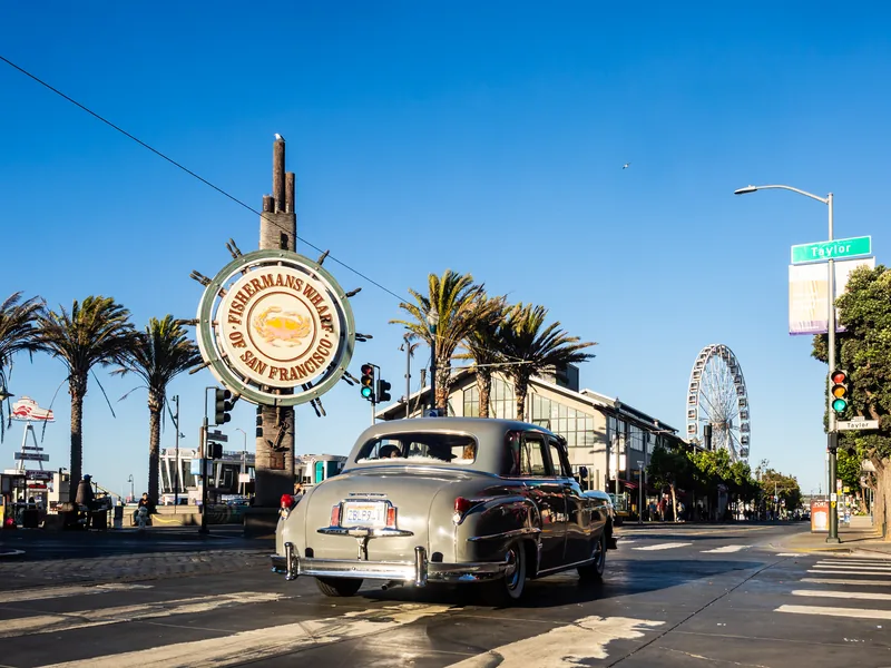 A vintage car passing the Fisherman's Wharf sign, with palm trees and a Ferris wheel.