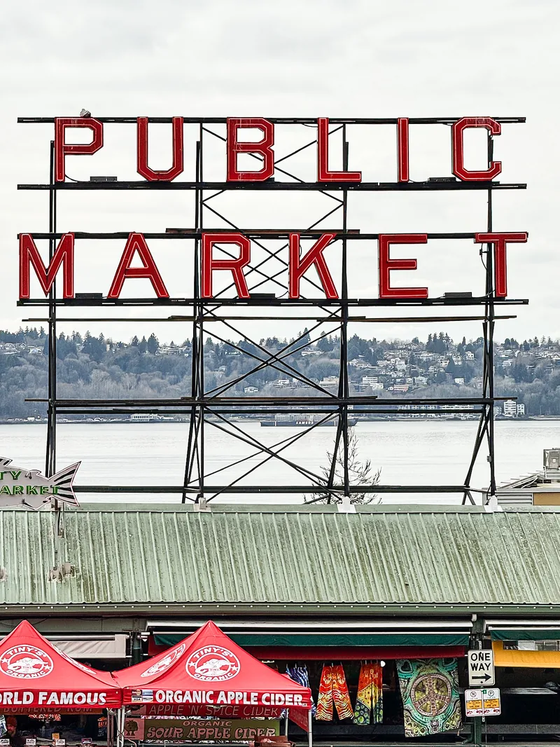 The Public Market sign on its steel frame above red umbrellas, with Elliott Bay behind.