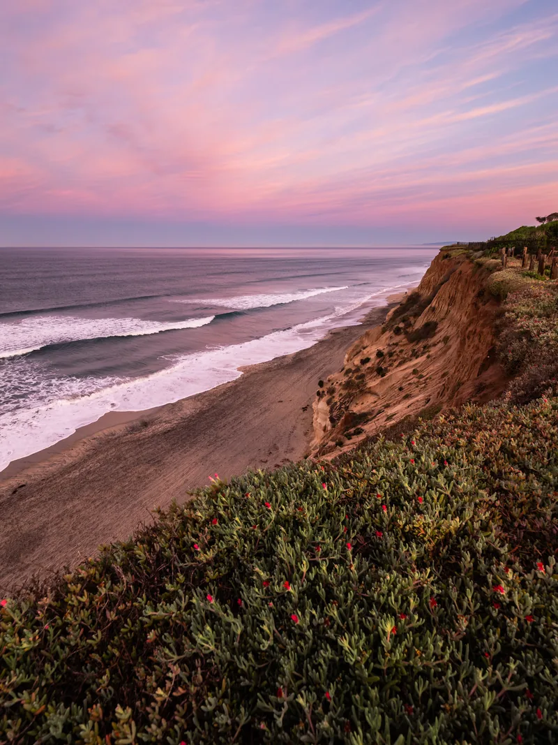 Waves washing up below a crumbling bluff under pink clouds.