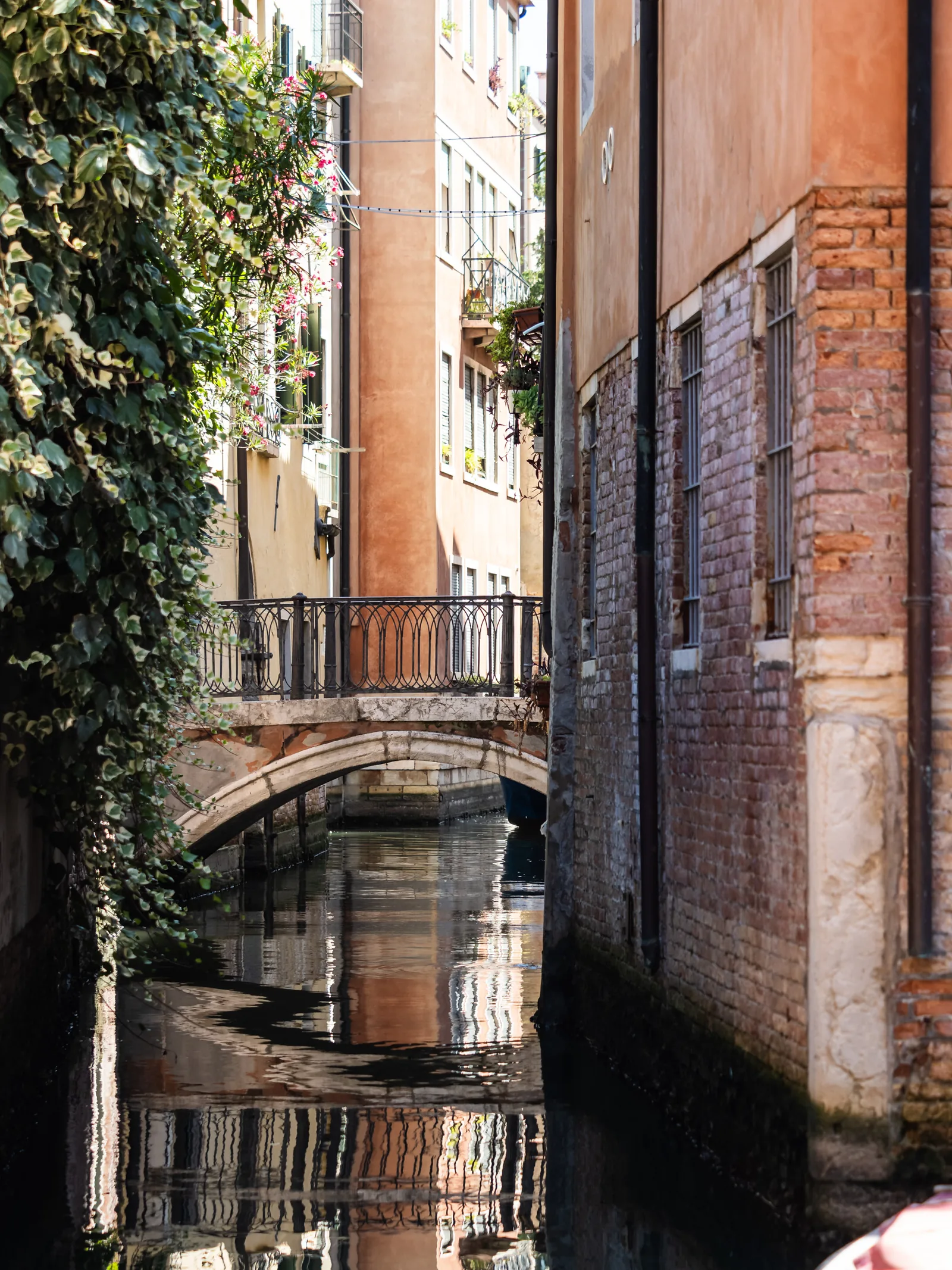 A narrow canal between old brick walls, with a small bridge and ivy reflected in the water.