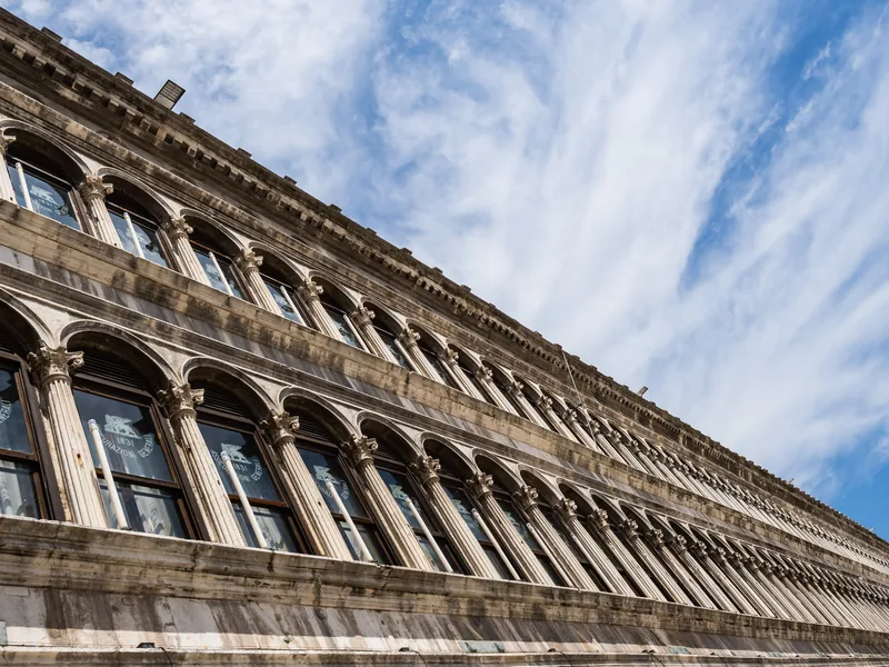A long arcaded façade on St Mark's Square, seen at a steep angle against wispy clouds.