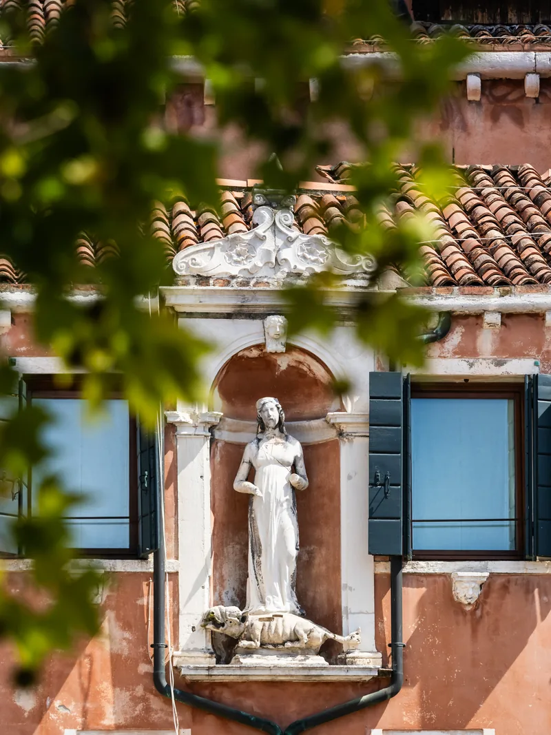 A white statue in a niche between shuttered windows on a terracotta wall, partly framed by leaves.
