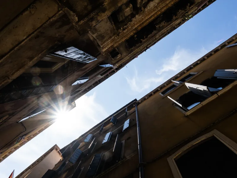 The sun bursting between two leaning building façades seen from below in a narrow street.