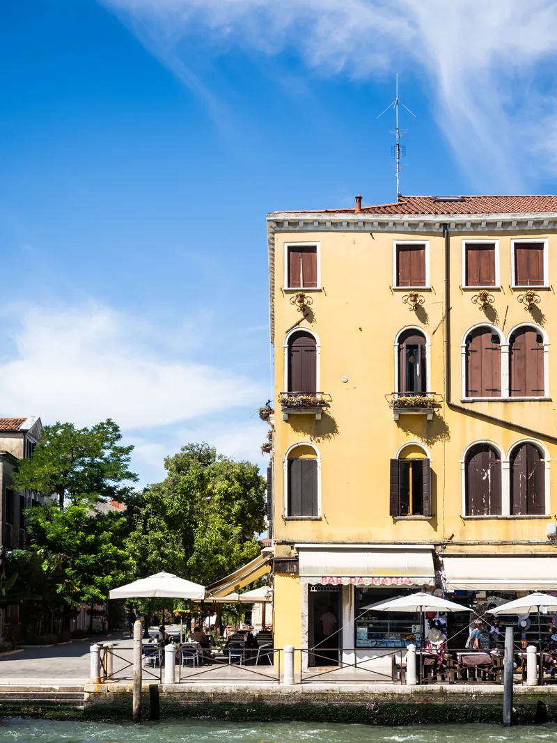 A yellow building with arched windows above café umbrellas at the water's edge.
