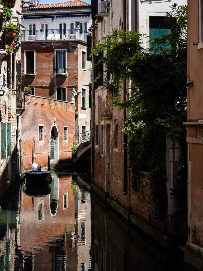 A shaded canal between old brick walls, a small boat tied up and greenery overhead.
