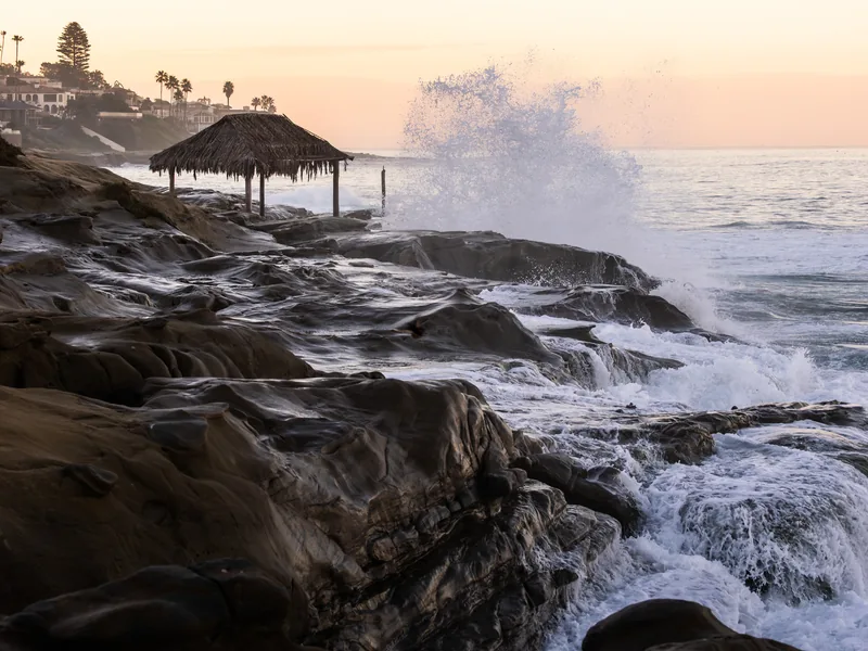 The Windansea palm-frond shack on the rocks as waves spray behind it.